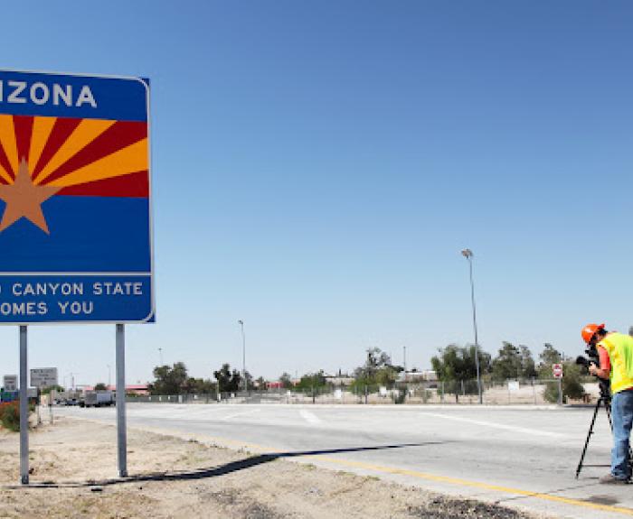 Arizona state welcome sign by a highway, with a worker in a safety vest standing nearby on the roadside.