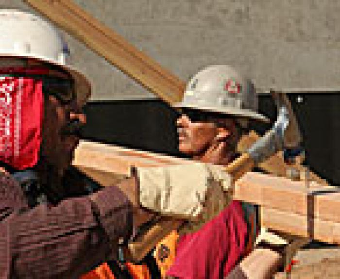 Two construction workers wearing hard hats and gloves hammer wooden beams at a building site.