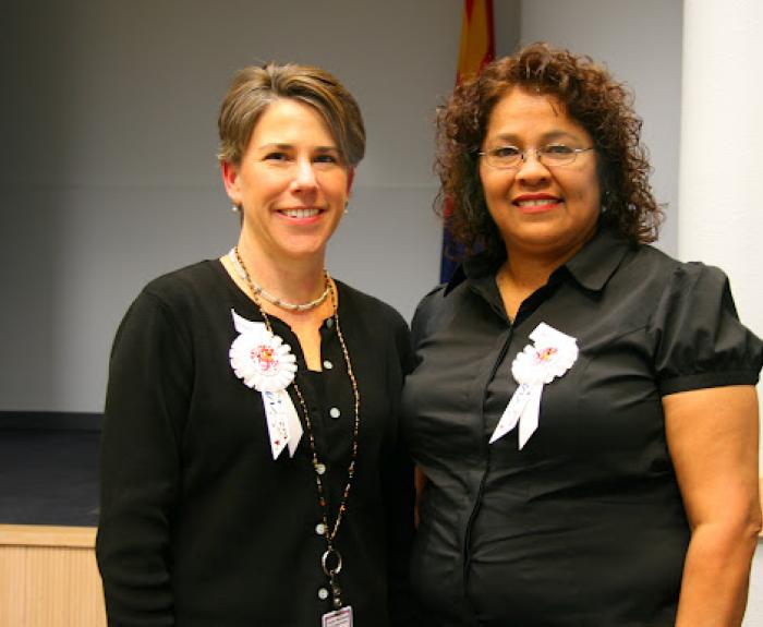 Two women stand side by side indoors, both wearing black tops and white ribbon badges, smiling at the camera.