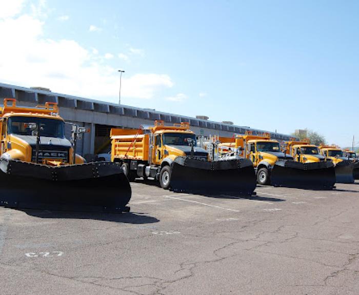 A row of yellow snow plow trucks are parked in a lot under a clear blue sky.