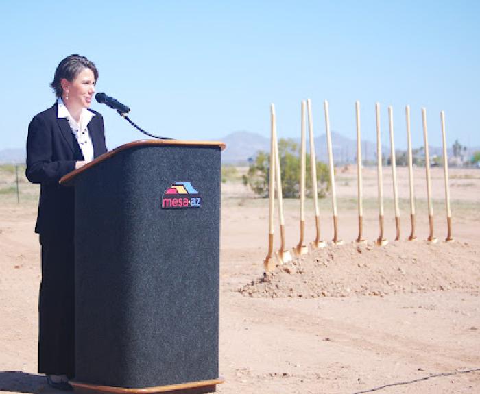 A woman speaks at a podium outdoors next to several golden shovels stuck in the dirt.