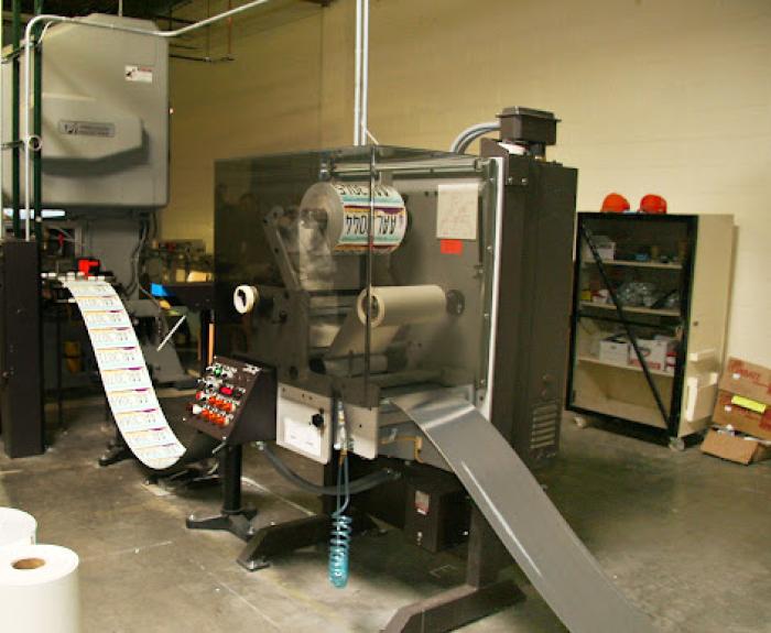 A license plate printing machine is producing a continuous roll of printed license plates from a sheet of steel. Shelves and boxes are visible in the background.