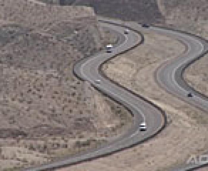 A winding highway curves through a dry, hilly desert landscape with several vehicles traveling in both directions.