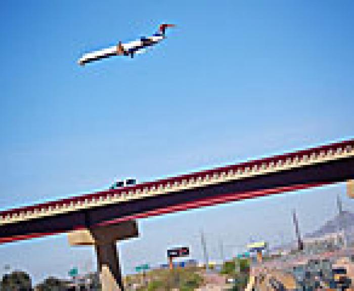 A commercial airplane flies low over a highway overpass with cars, against a clear blue sky and distant mountains in the background.
