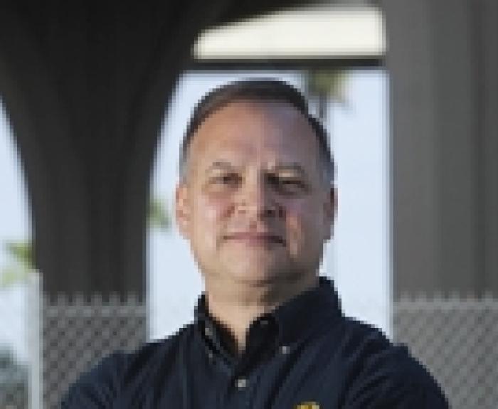 A man stands outdoors with arms crossed, wearing a black button-up shirt with a yellow logo, in front of a chain-link fence and large architectural columns.