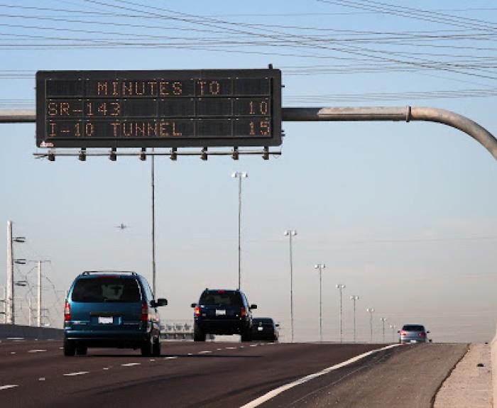 Cars driving on a highway under an electronic sign displaying travel times: “MINUTES TO SR-143: 10, I-10 TUNNEL: 15,” with a clear sky and power lines overhead.