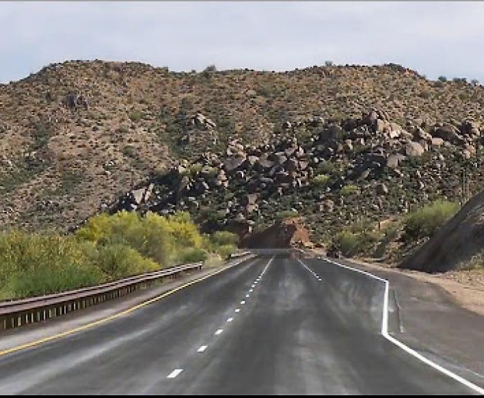 A two-lane highway leads toward a rocky hill covered with sparse desert vegetation under a partly cloudy sky. There are guardrails and shoulder areas on each side.