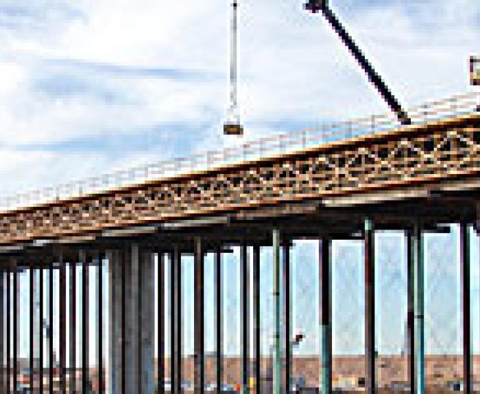 A crane lifts construction materials onto a long, elevated bridge supported by tall pillars, with a partly cloudy sky in the background.