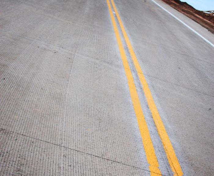 A concrete road with double yellow lines runs uphill under a blue sky, with streetlights and dirt embankments on either side.