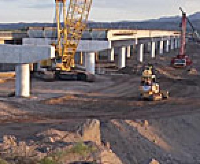 Construction equipment working on a bridge with unfinished concrete supports and dirt piles nearby.