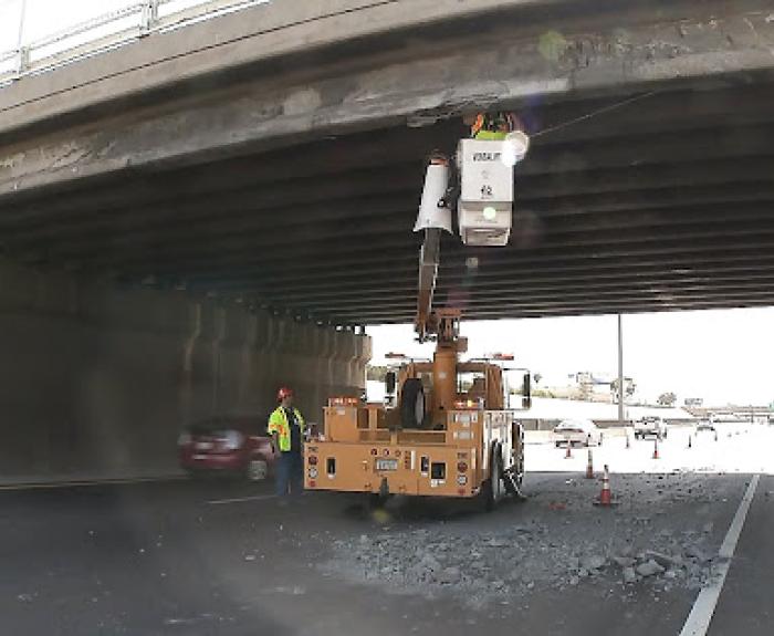 Workers use a bucket truck to inspect and repair damage under a highway overpass while debris is scattered on the closed roadway below.