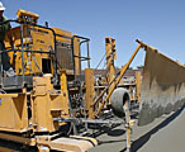 A construction worker operates a large yellow paving machine that is spreading and smoothing fresh concrete on a road under a clear blue sky.