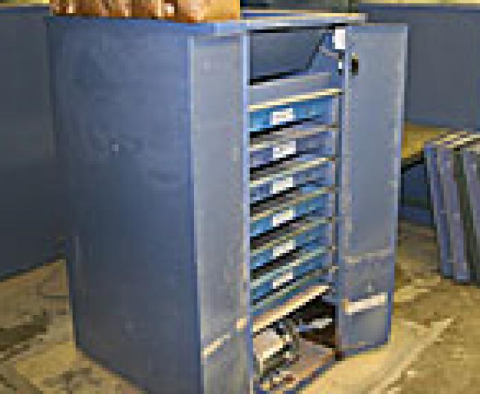 A blue metal storage cabinet with multiple open drawers stands on a concrete floor in an industrial setting. Several flat cases are propped against the wall nearby.