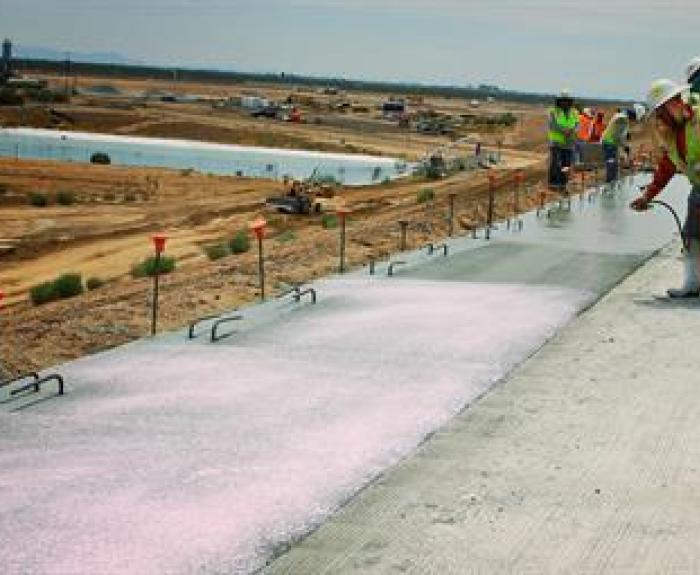 Workers in safety vests and helmets apply curing compound to a concrete surface at a large outdoor construction site with earthmoving equipment in the background.