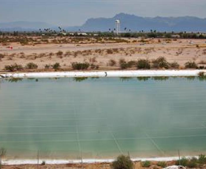 A large, rectangular, man-made pond lined with white material sits in a desert landscape with sparse vegetation and distant mountains under a cloudy sky.