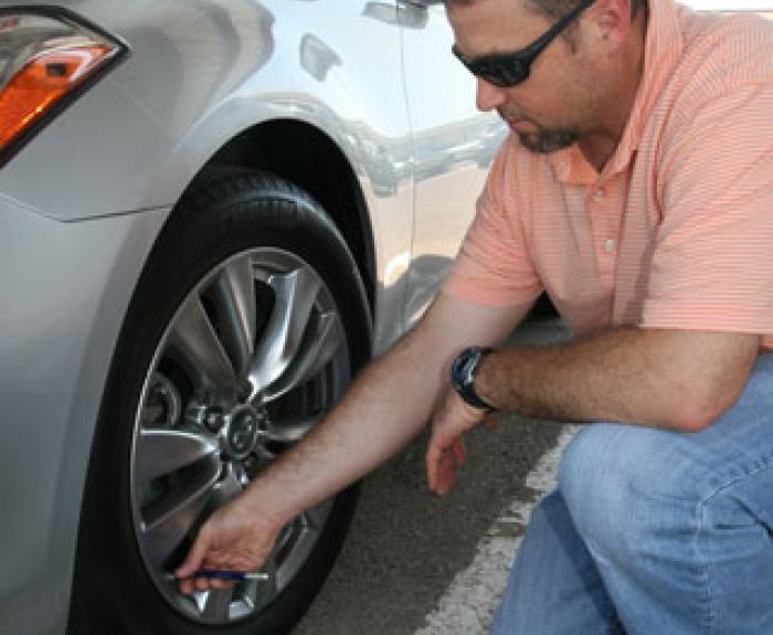A man wearing sunglasses and a striped polo shirt crouches to check the tire pressure of a parked silver car using a tire pressure gauge.