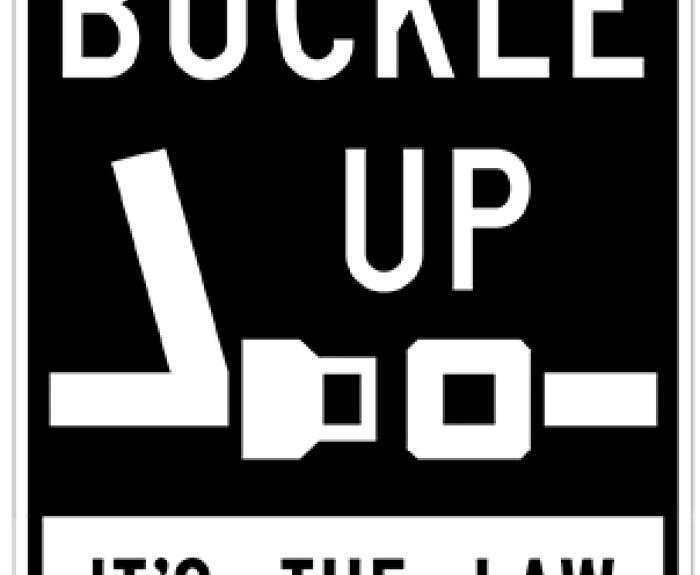 Black and white sign showing a seatbelt icon with the words “Buckle Up, It’s the Law.”.