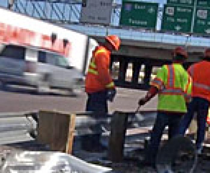 Three construction workers in orange safety vests stand beside a metal guardrail on a highway, with a truck and road signs visible in the background.
