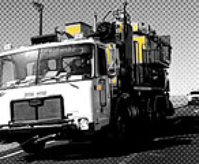 A large road maintenance vehicle travels on a highway; image is mostly black and white with yellow highlights.