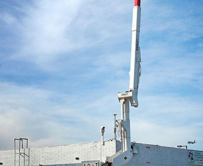 A yellow utility truck with an extended aerial lift holds a worker in a bucket raised high near a building under a partly cloudy sky.