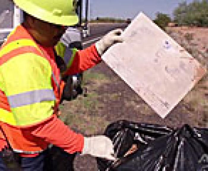 A worker in a safety vest and helmet picks up litter and places it in a black trash bag by the roadside.