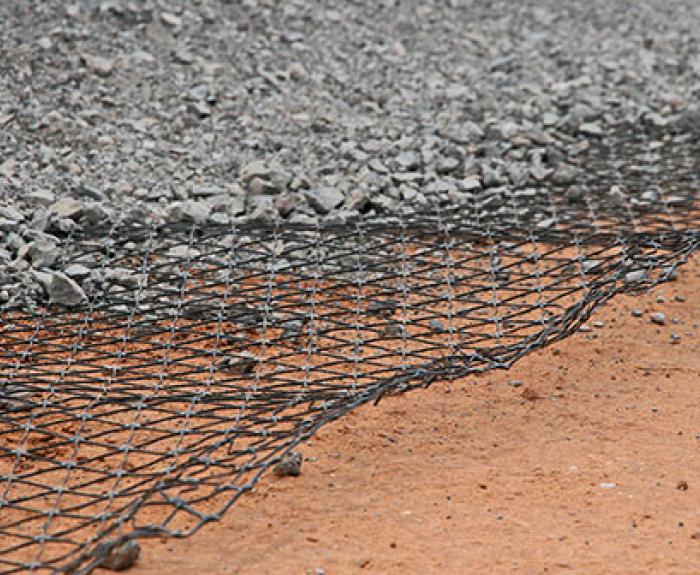 A layer of geogrid mesh lies between compacted gravel above and reddish-brown soil below, illustrating ground reinforcement in construction.