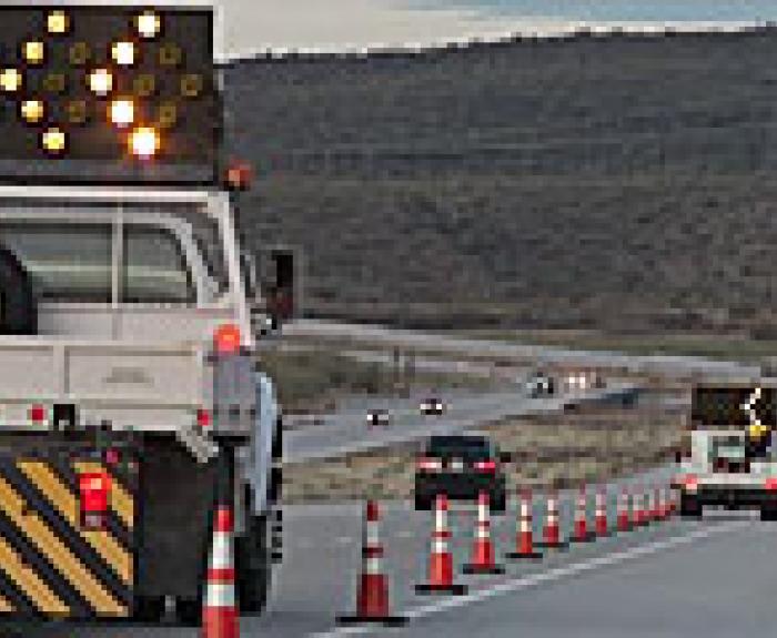 Two trucks with arrow boards and striping guide traffic through a construction zone marked by orange cones on a highway; several cars drive nearby.