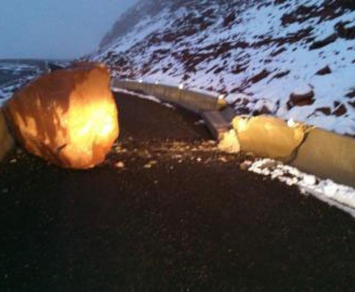 A large boulder blocks a snow-covered mountain road, breaking through a concrete barrier; headlights illuminate the scene in low light.
