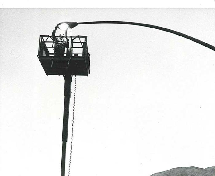 A person stands on the roadside watching a worker in a lift repair a streetlight above a curved road.