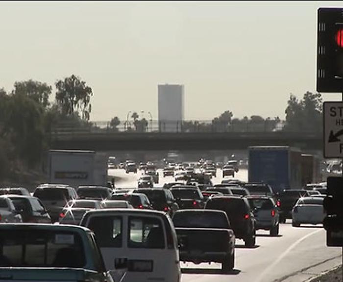 Heavy traffic fills multiple lanes on a freeway under hazy daylight, with a red traffic light and “Stop Here On Red” sign visible in the foreground.
