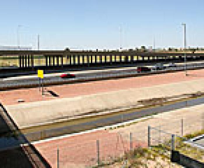 A highway overpass with several cars driving, crossing above a dry concrete drainage canal on a clear, sunny day. A green road sign is visible in the distance.