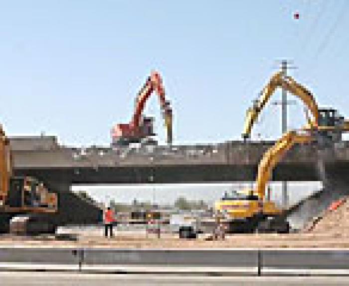 Excavators demolishing a bridge while construction workers oversee the site on a sunny day.