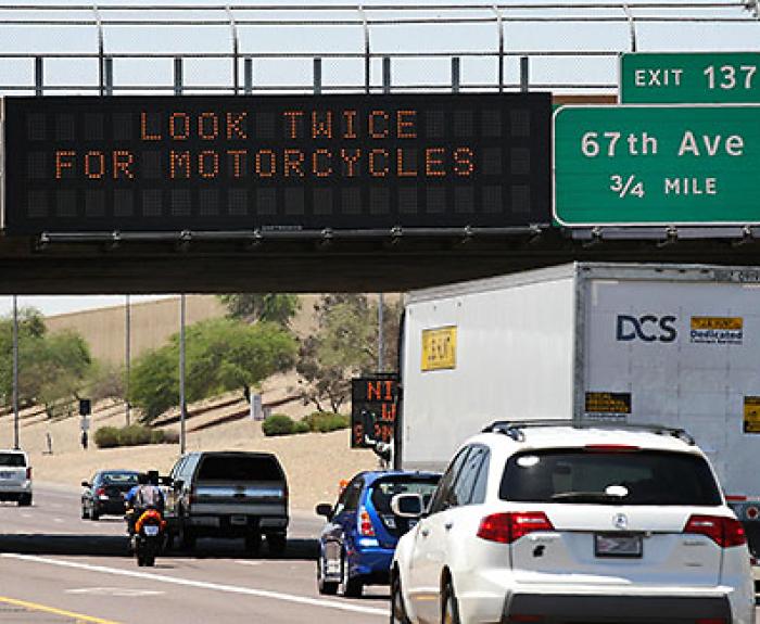 Cars and a motorcycle drive on a highway under a digital sign that reads, LOOK TWICE FOR MOTORCYCLES; exit sign for 67th Ave is visible.