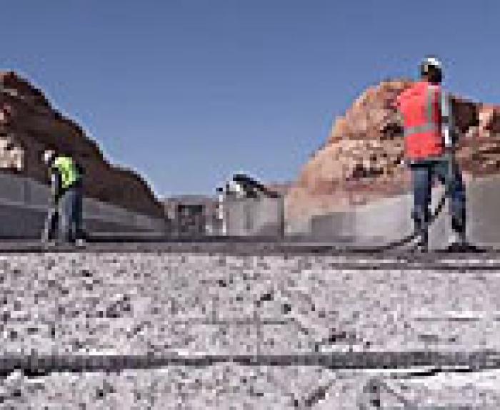 Two construction workers in safety gear pour and spread concrete on a bridge, with rocky hills and clear blue sky in the background.