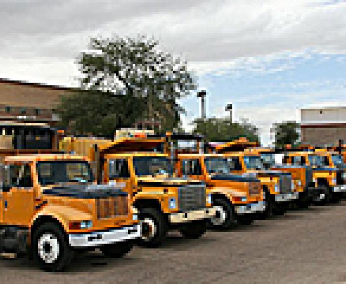 A row of yellow trucks in a parking lot near buildings and trees under a cloudy sky.