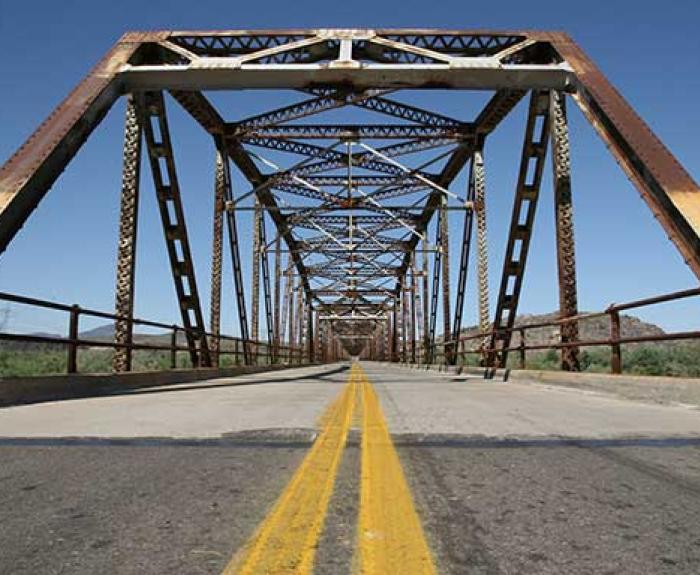 A view down the center of an old, rusted steel truss bridge with a double yellow line on the road, under a clear blue sky.
