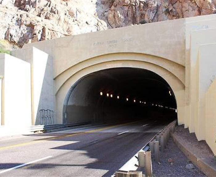 A concrete tunnel entrance set into a rocky hillside, with lights lining the inside and a two-lane road leading into the tunnel.