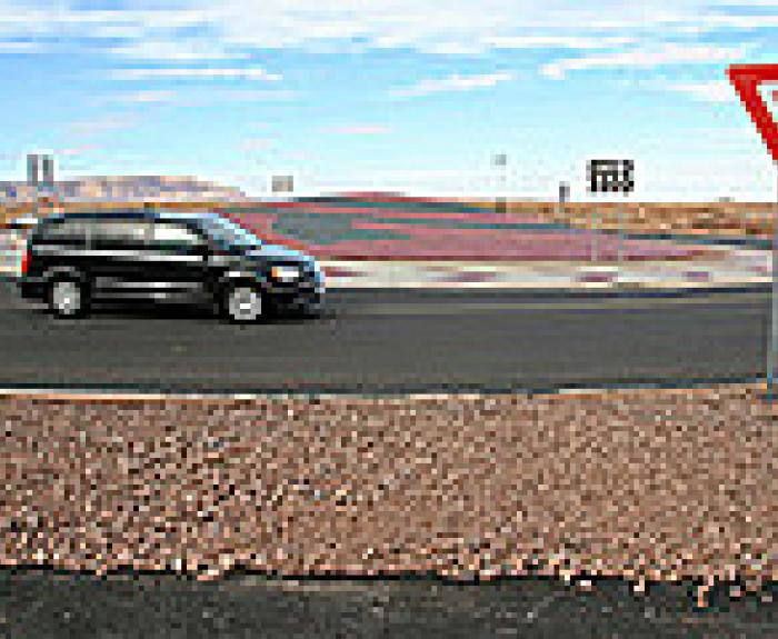 Black minivan enters a roundabout near a yield sign on a clear day with a desert background.