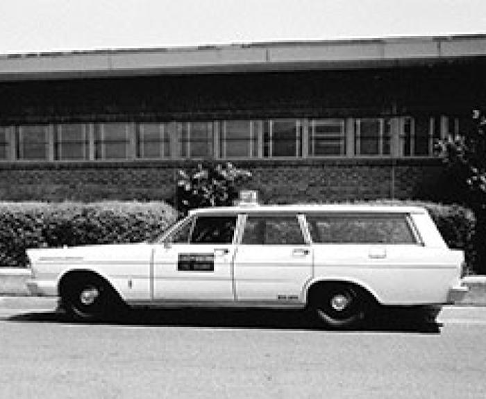 Black and white photo of a vintage station wagon taxi parked in front of a low building with bushes.