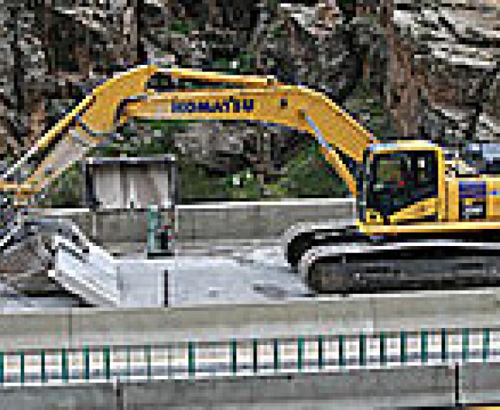A yellow excavator works beside a concrete barrier on a road in a rocky, mountainous area.