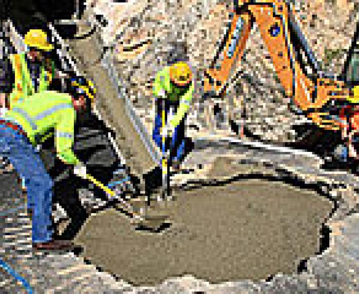 Workers in safety gear pour and spread concrete into a large hole at a construction site with machinery nearby.