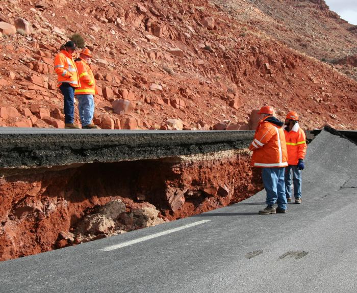 Three workers in orange jackets inspect a road severely damaged and lifted by a landslide in a rocky area.