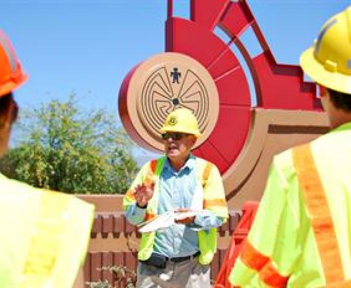 Three construction workers in safety gear talk near a large, red sculptural sign under a clear blue sky.