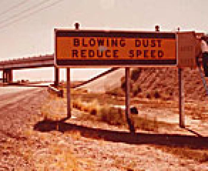 A person sits on a ladder next to a roadside sign reading BLOWING DUST REDUCE SPEED near a highway.