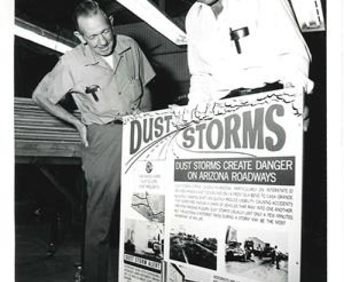 Two men examine a large poster warning about the dangers of dust storms on Arizona roadways.