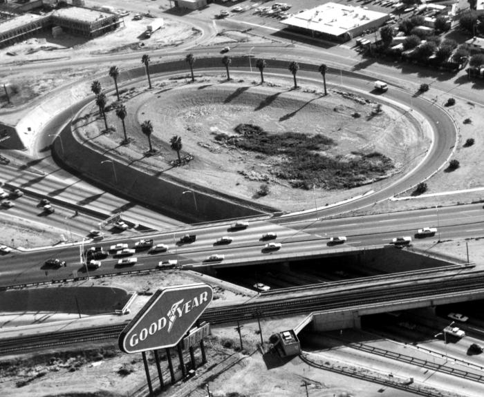 Aerial view of a freeway interchange with multiple cars, a central oval island lined with palm trees, and a large Goodyear sign beside railroad tracks in the foreground.
