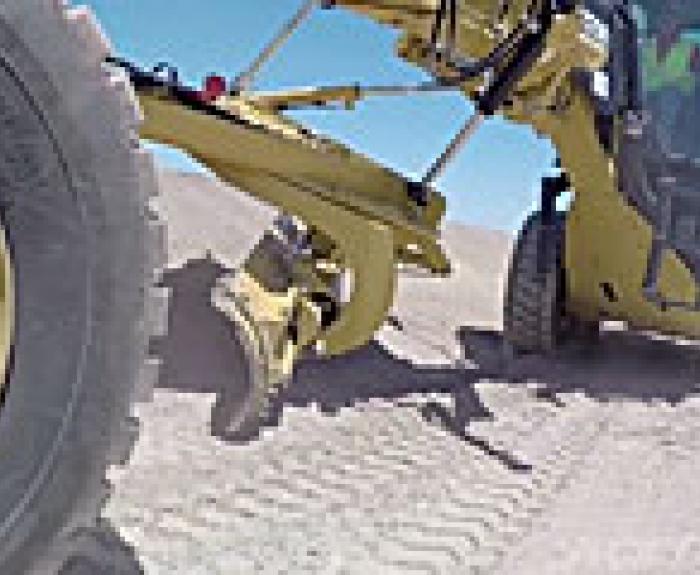 A close-up view of a yellow construction grader smoothing a dirt road under a clear blue sky, with visible tire tracks and part of the operator’s cabin.