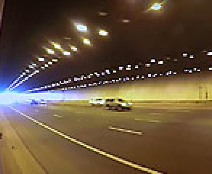 Cars drive through a wide, well-lit tunnel with multiple lanes; bright light is visible at the tunnels distant exit.