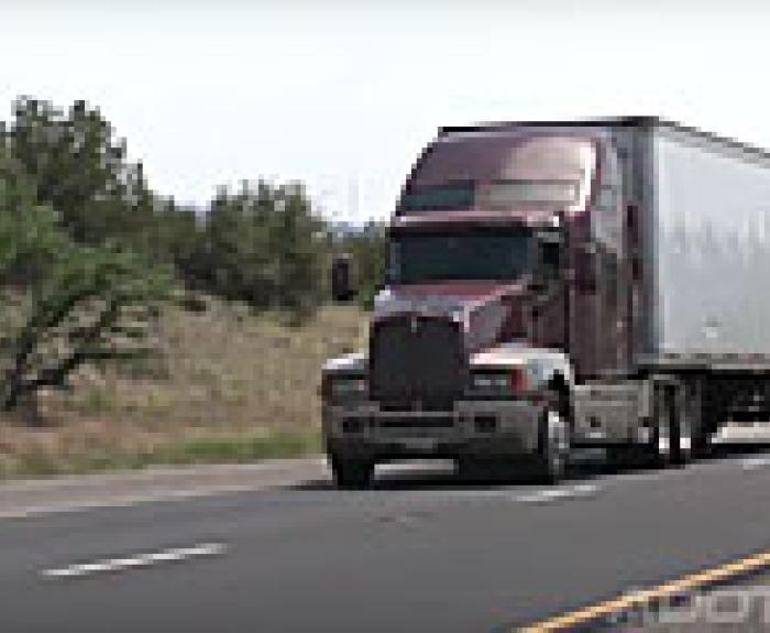 A red semi-truck with a white trailer driving on a highway surrounded by trees and grass.