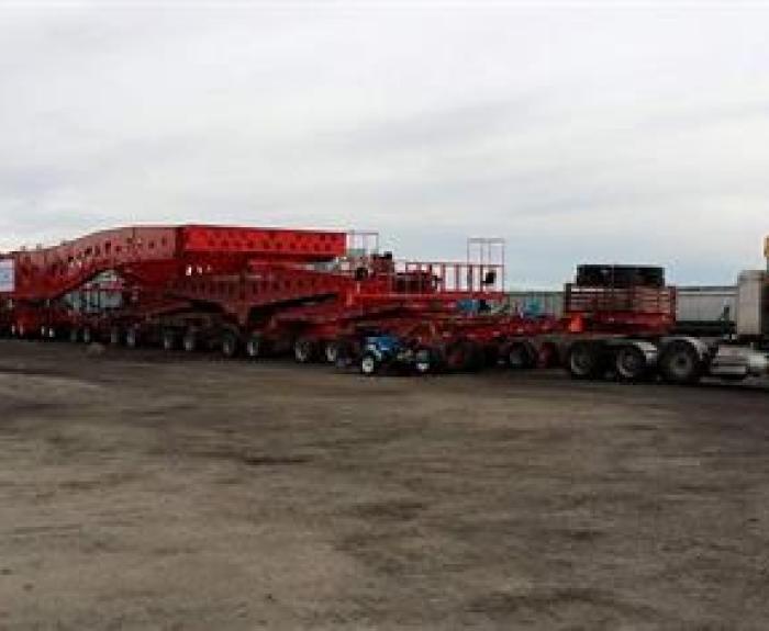 A large red modular trailer with multiple axles is connected to a red semi-truck, parked on a wide gravel lot under an overcast sky.